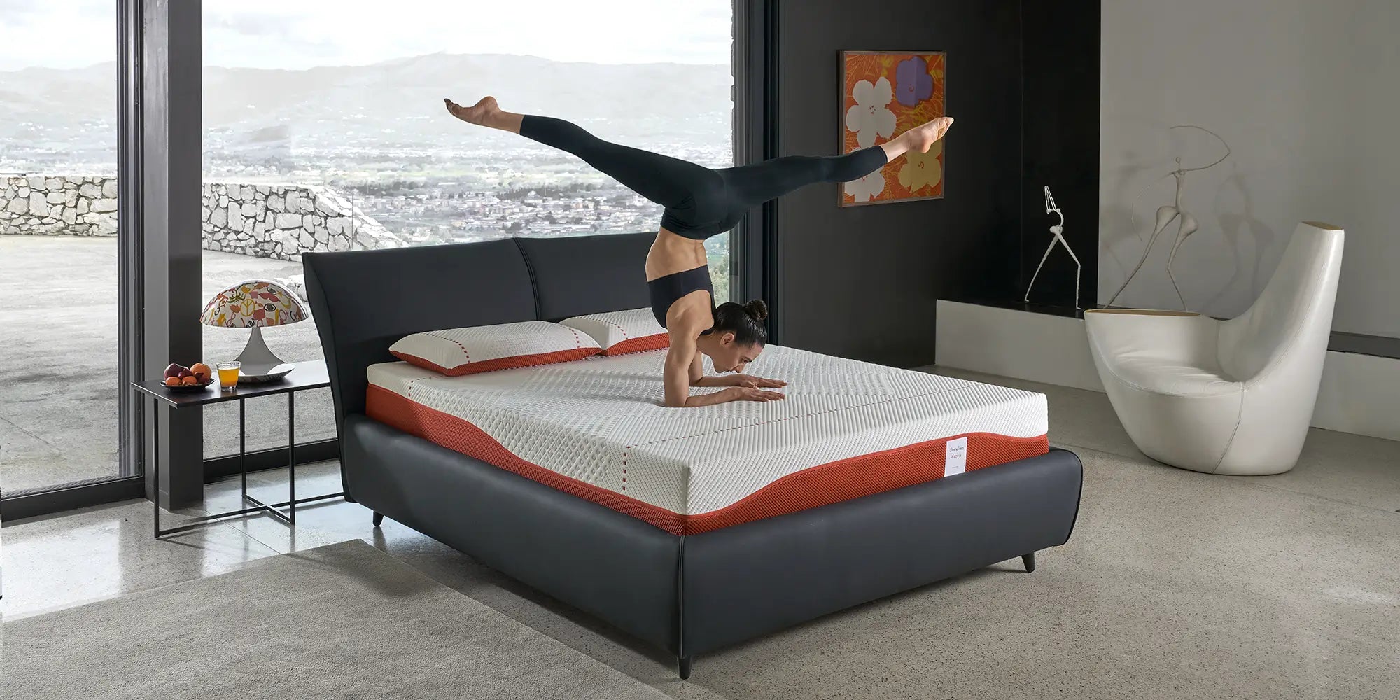 An athlete performing a handstand on a Dorelan ReActive mattress with a black bed frame, set in a modern bedroom with a panoramic window view.
