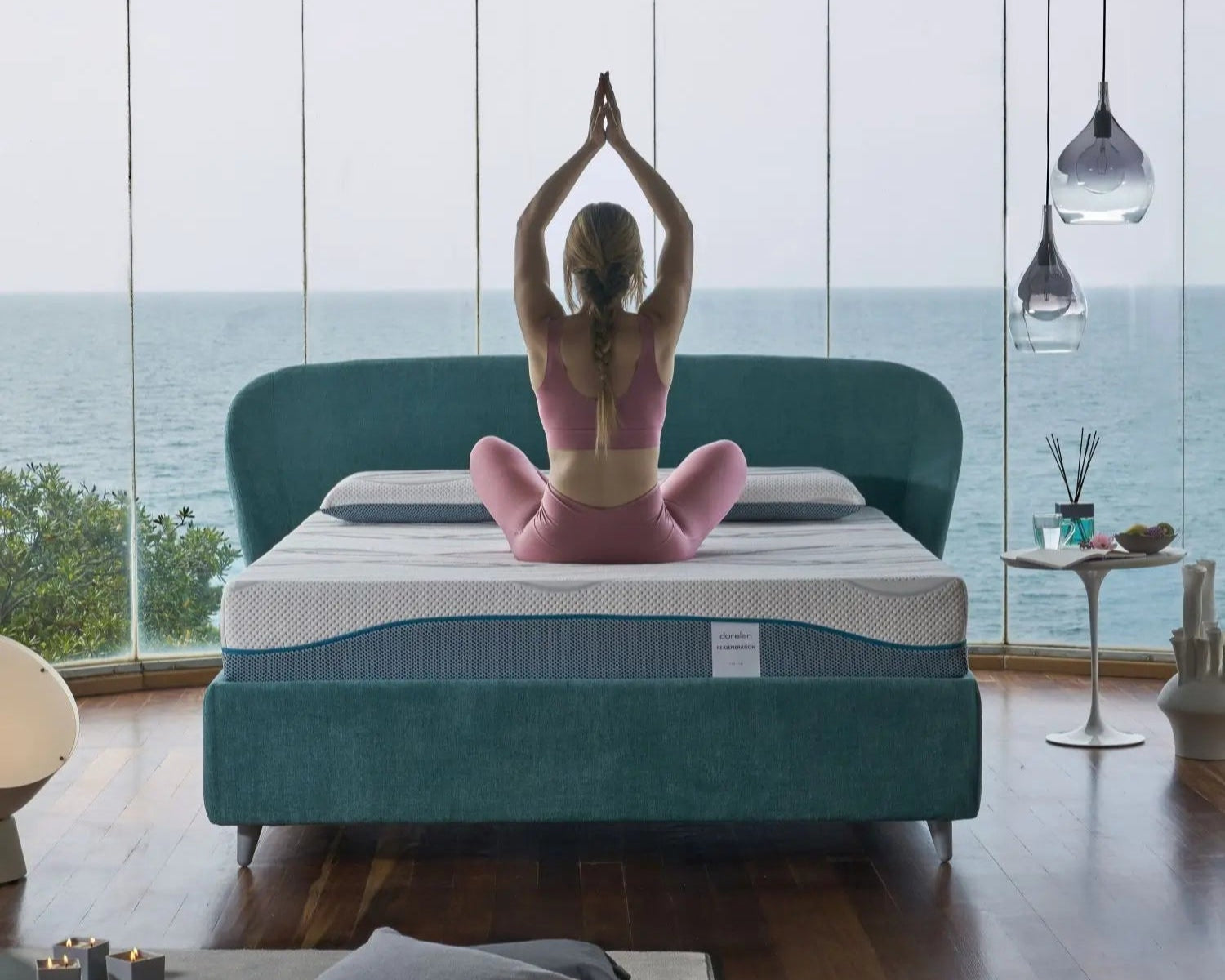 A woman practicing yoga on a Dorelan Re:Generation mattress with a teal bed frame, in a tranquil room overlooking the sea.