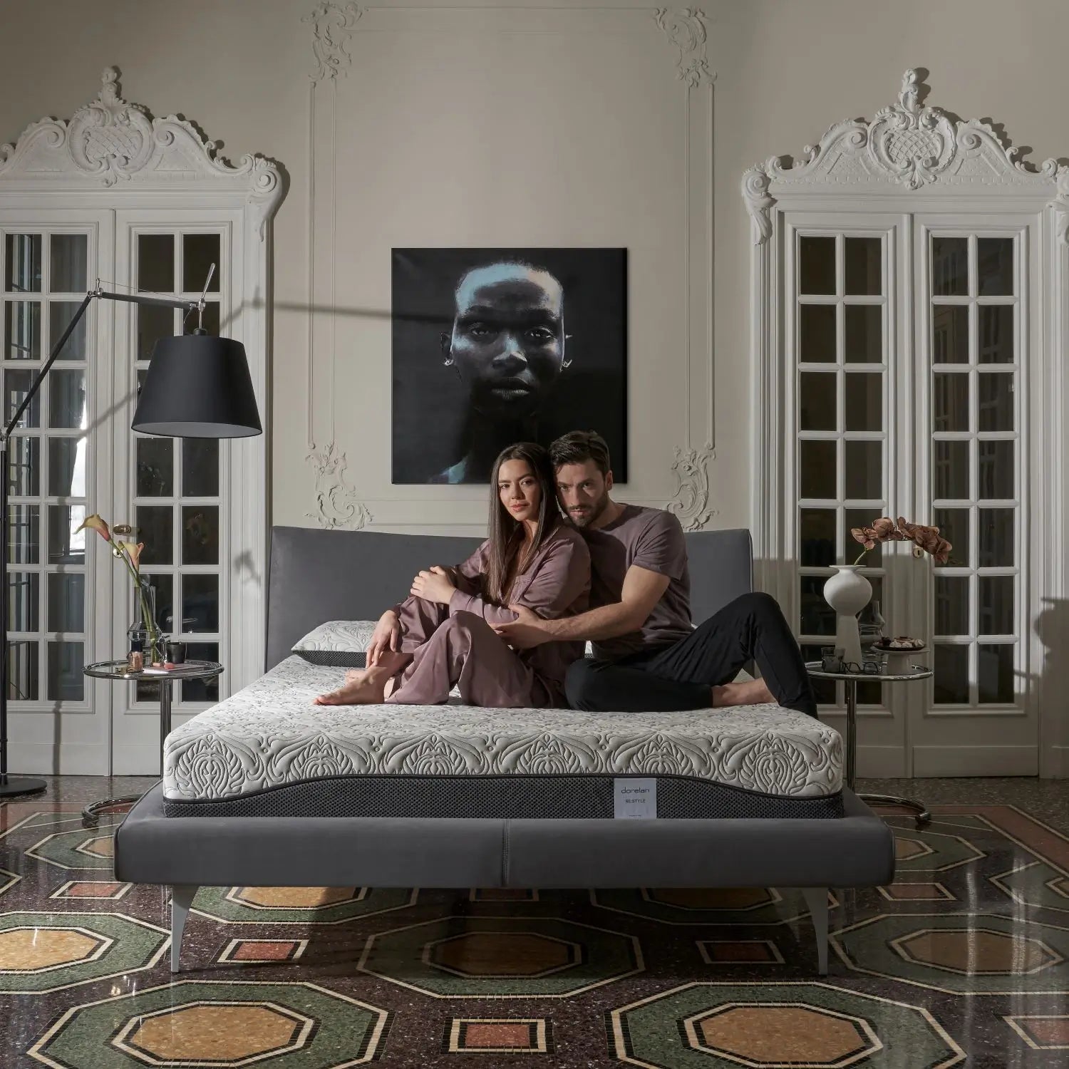 A couple sitting on a Dorelan Re:Style mattress with an elegant gray bed frame, in a classic room with ornate decor and soft lighting.