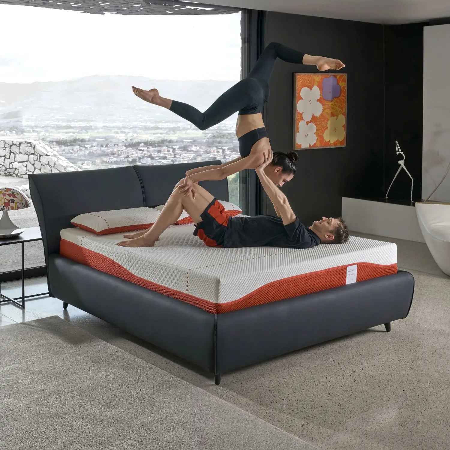 A couple performing an acrobatic pose on a Dorelan Re:Active mattress with a black bed frame, set in a modern bedroom with a panoramic view.