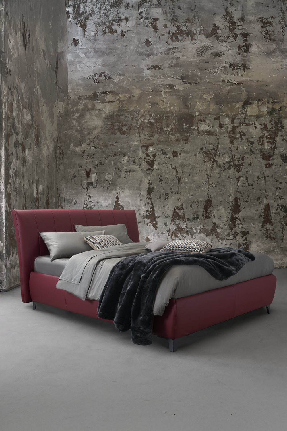 Letto imbottito bordeaux con testiera trapuntata, biancheria grigia e plaid in eco pelliccia, su sfondo materico