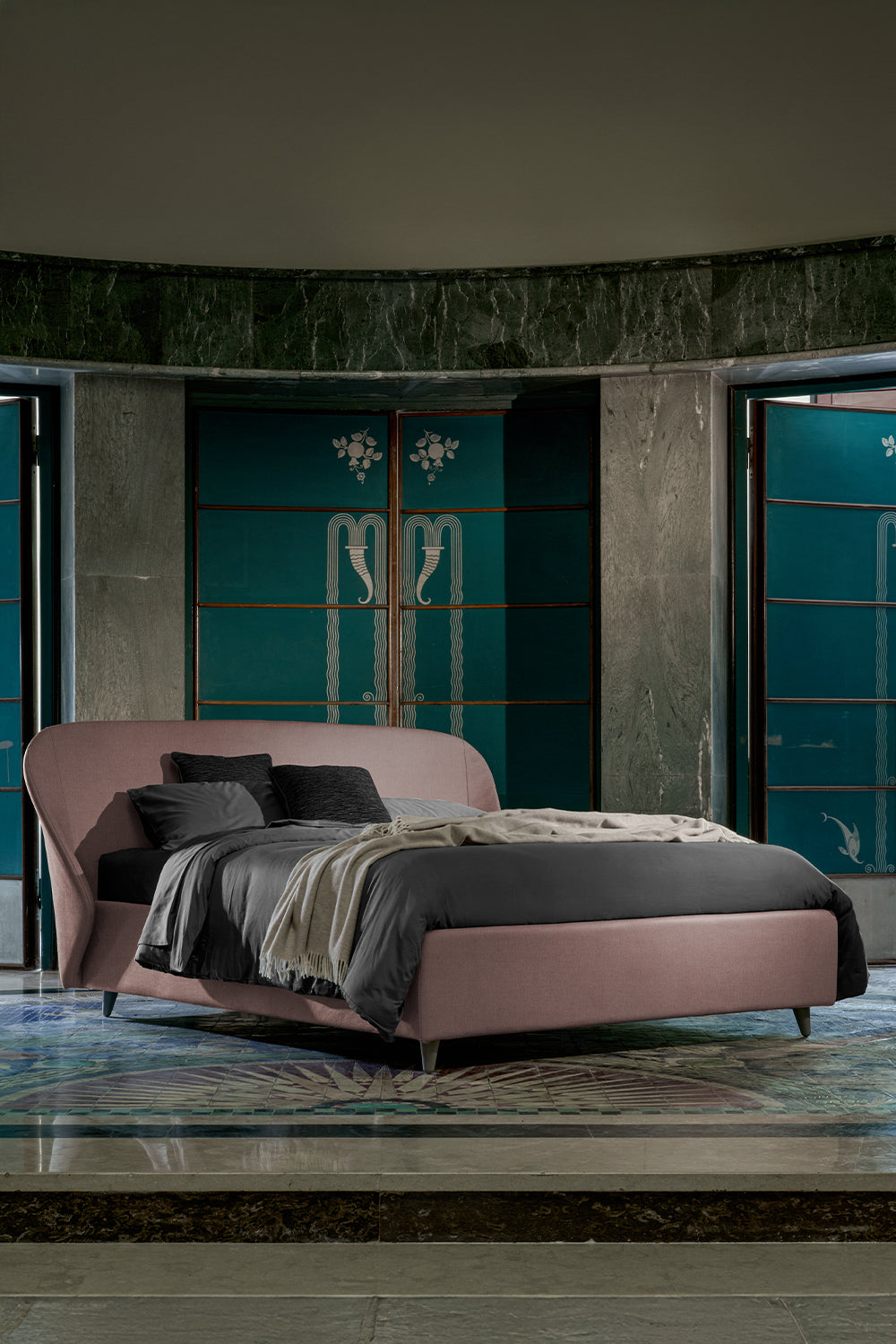 Letto rosa cipria con coperta grigia e cuscini neri, posizionato in una stanza elegante con pareti verdi e decoro Art Deco