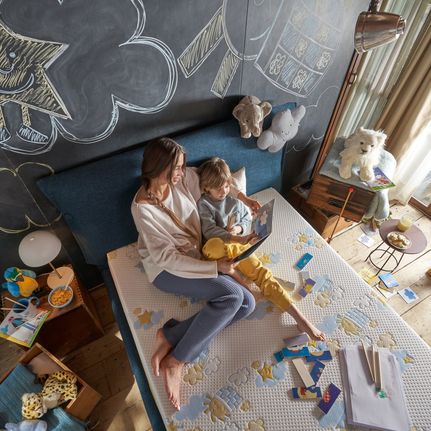 mamma e figlio leggono la favola della buona notte su un materasso con i disegni di nicoletta costa, in una cameretta con giochi e peluche