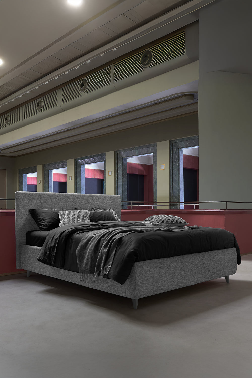 Letto matrimoniale grigio con lenzuola nere e cuscini in tinta, ambientato in uno spazio architettonico contemporaneo con colonne e vetrate